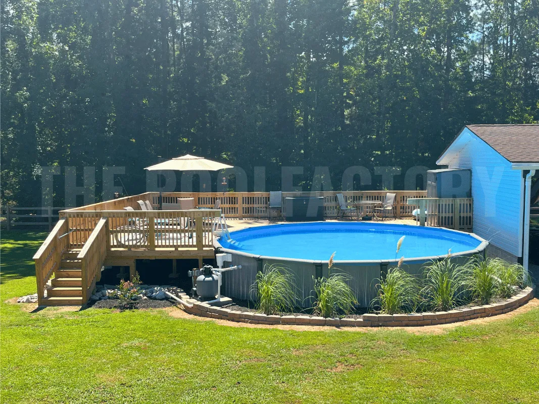 Round above ground pool surrounded by deck, lush plants, and small retaining wall