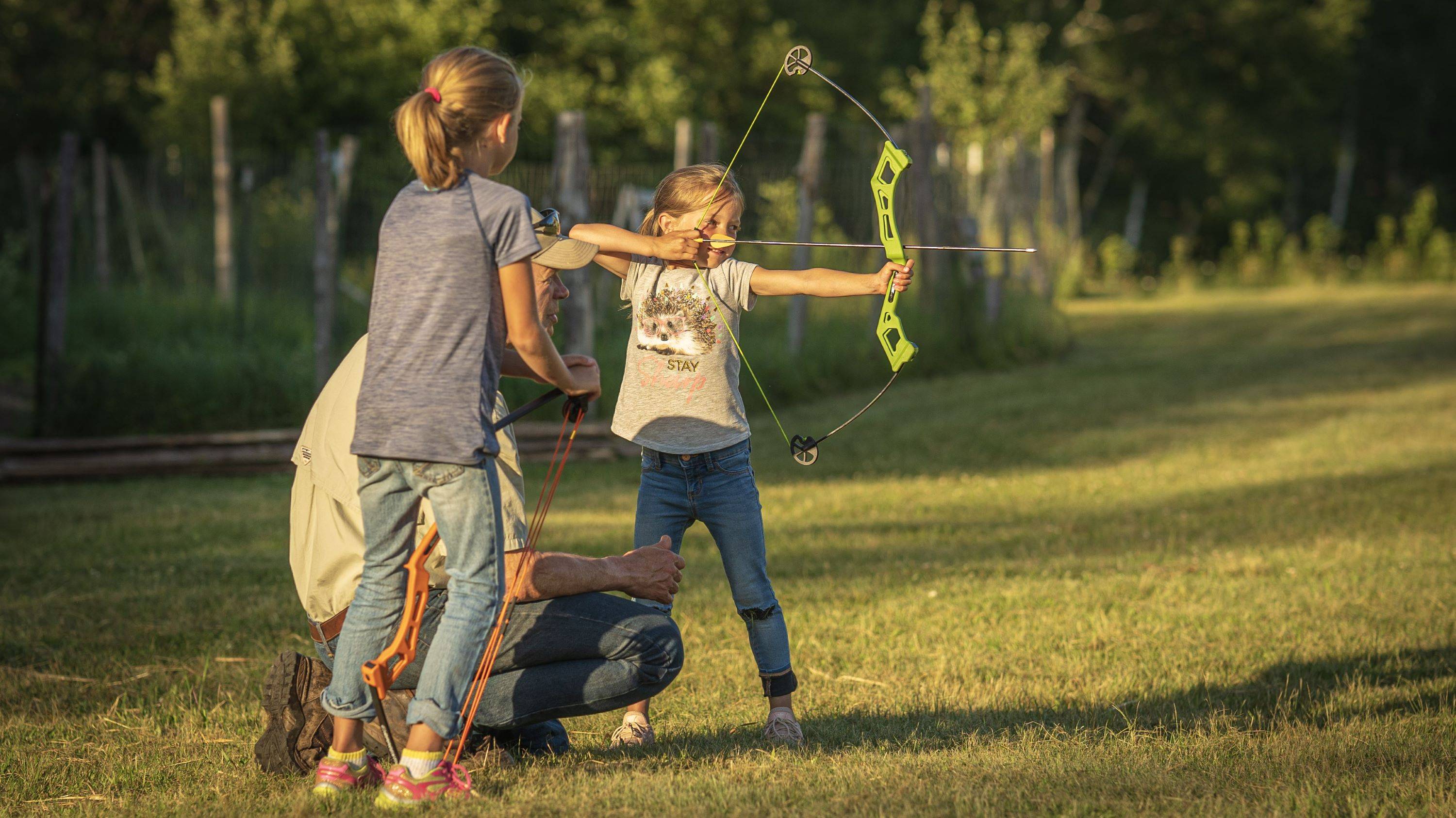 Two girls standing next to their grandpa while one of them shoots the Bear Archery Valiant youth bow