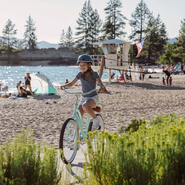 A child rides a light blue Chatham 24" Kids' Beach Cruiser Bike on a sandy beach, with trees and a lifeguard station in the background, enjoying a sunny day.