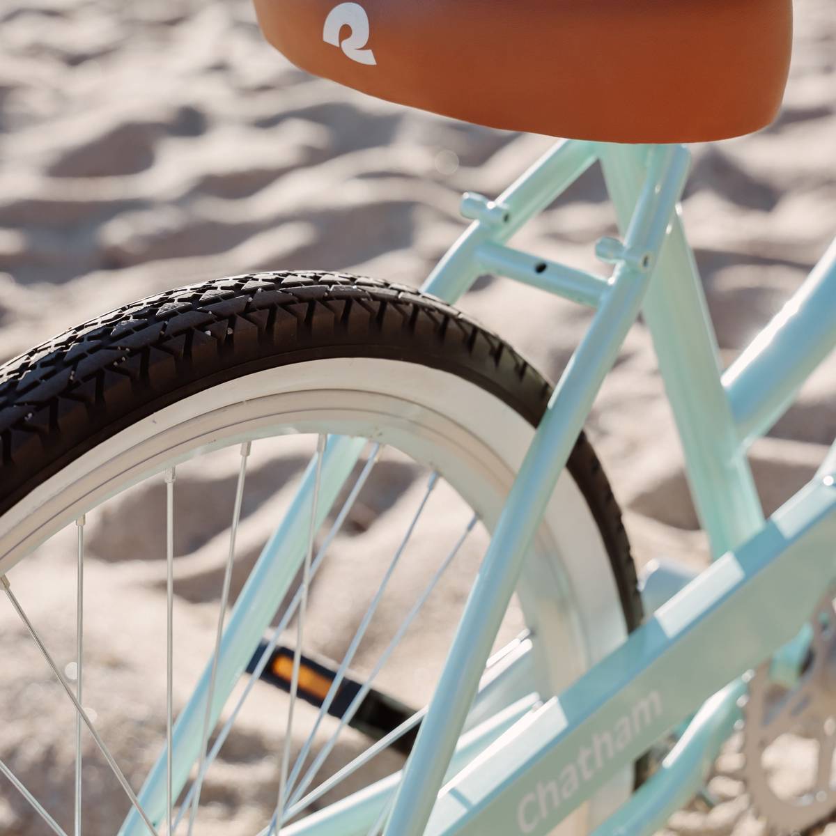 Close-up of a light blue Chatham 24" Kids' Beach Cruiser Bike with a brown seat, showcasing the textured tire and sandy background.
