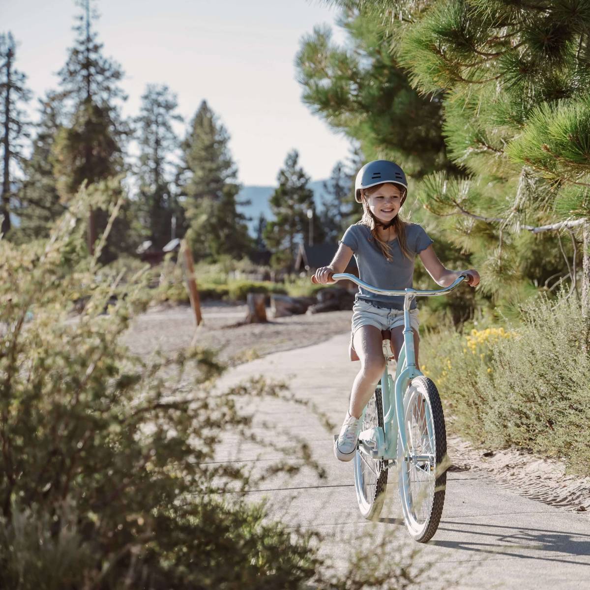 A girl rides a light blue Chatham 24" Kids' Beach Cruiser Bike along a serene, sunlit path surrounded by trees and greenery in a picturesque outdoor setting.