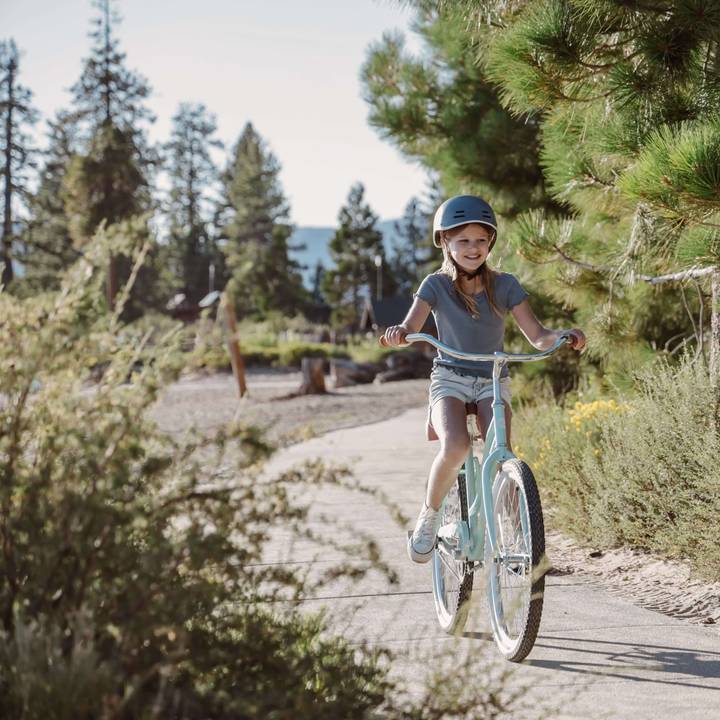 A girl rides a light blue Chatham 24" Kids' Beach Cruiser Bike along a serene, sunlit path surrounded by trees and greenery in a picturesque outdoor setting.