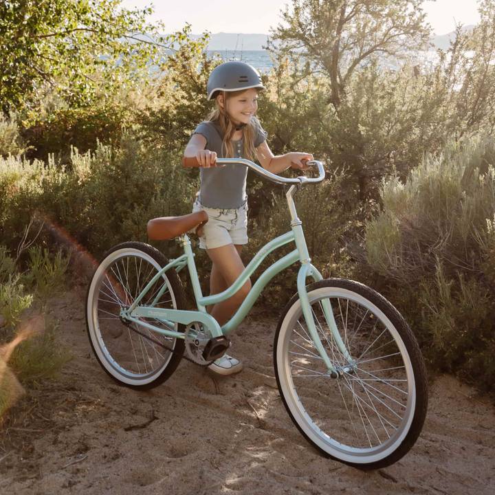 A child in a gray helmet rides an aqua Chatham 24" Kids' Beach Cruiser Bike on a sandy path surrounded by greenery, with a serene water view in the background.