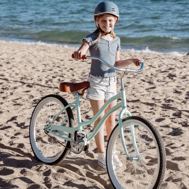 A child stands on a sandy beach, holding a vintage-style mint green Chatham 24" Kids' Beach Cruiser Bike, dressed in a light blue shirt and shorts with a helmet on.