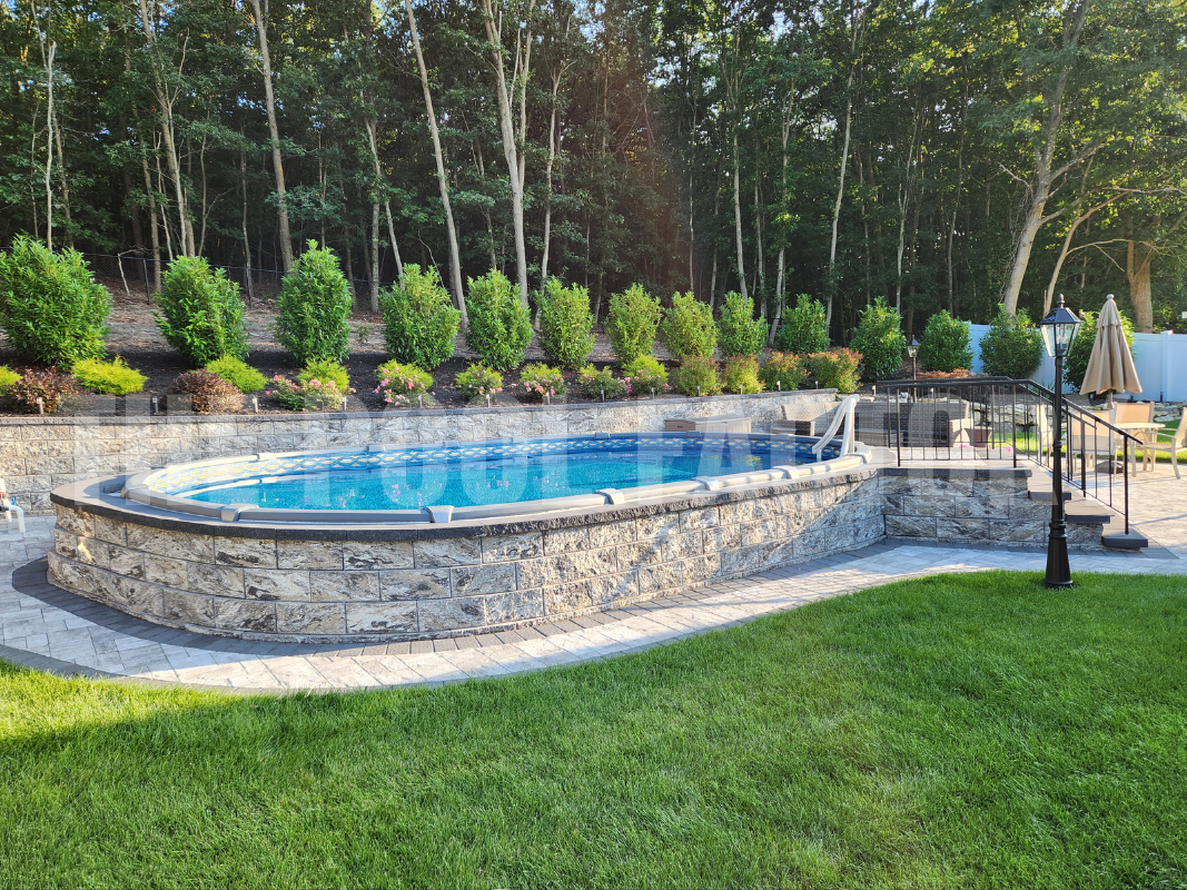 Semi-inground oval saltwater pool with stone patio, flowers, and shrubs