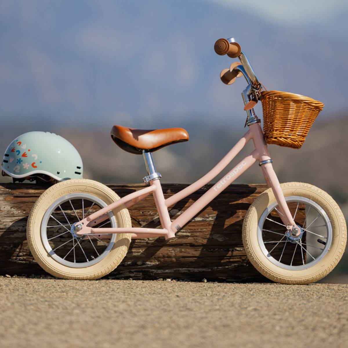 A pink Baby Beaumont 12" Kids' Balance Bike with a brown seat and basket, beside a light green helmet, against a scenic outdoor background.