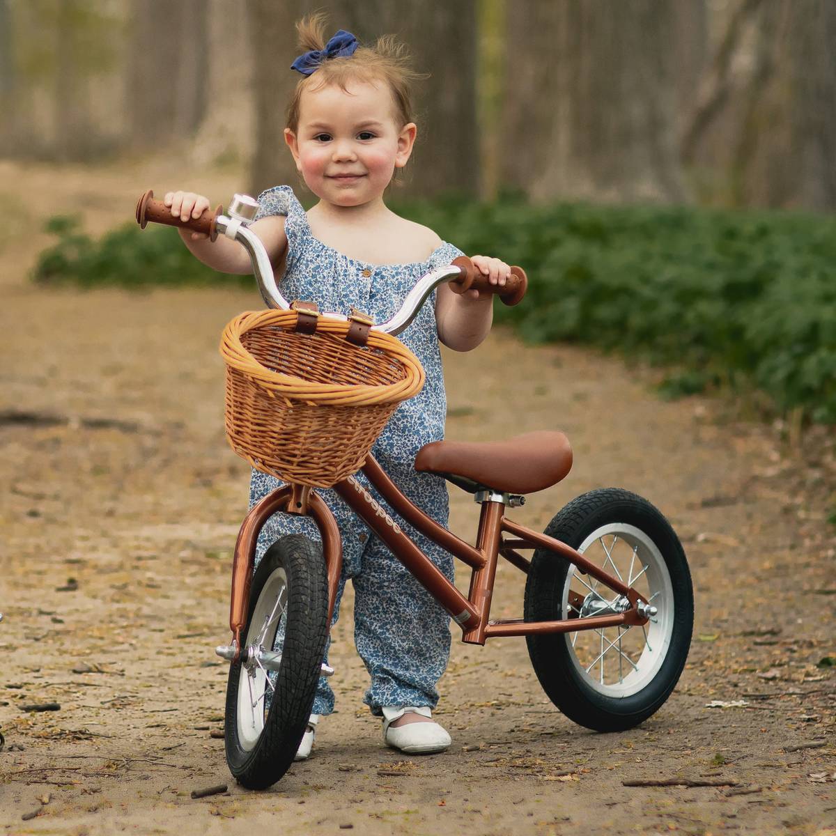 A child in blue jumper holds the handlebars of a rust colored Baby Beaumont 12" Kids' Balance Bike with a wicker basket, standing on a dirt path surrounded by greenery and trees.