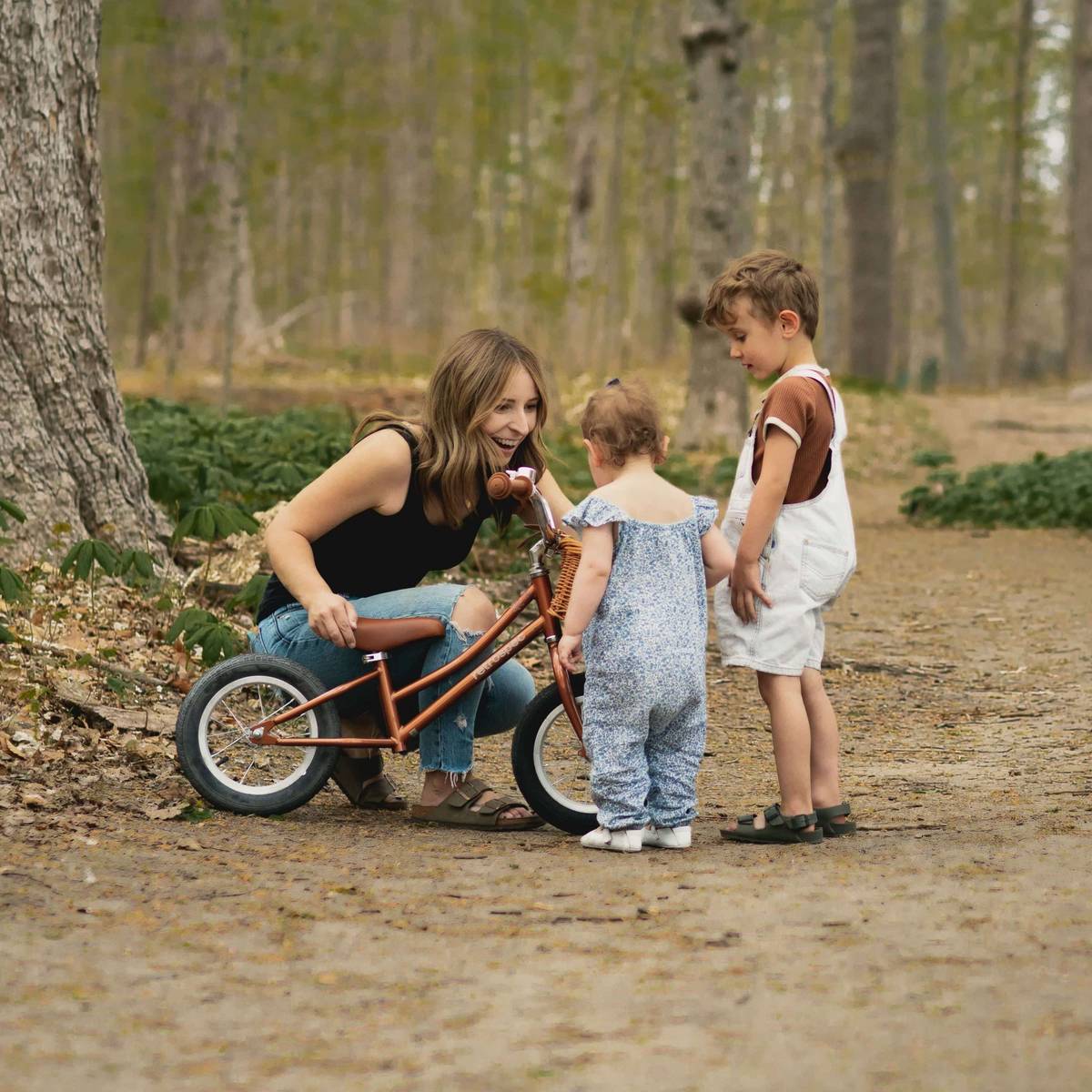 A woman crouches and smiles as two young children explore a copper-toned Retrospec balance bike on a wooded dirt path.