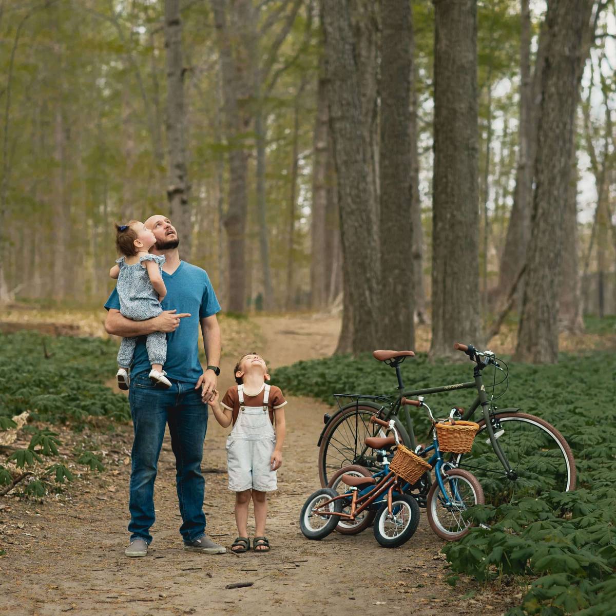 A father holds his toddler and walks with his young son on a wooded trail, with a lineup of Retrospec bikes parked alongside them.