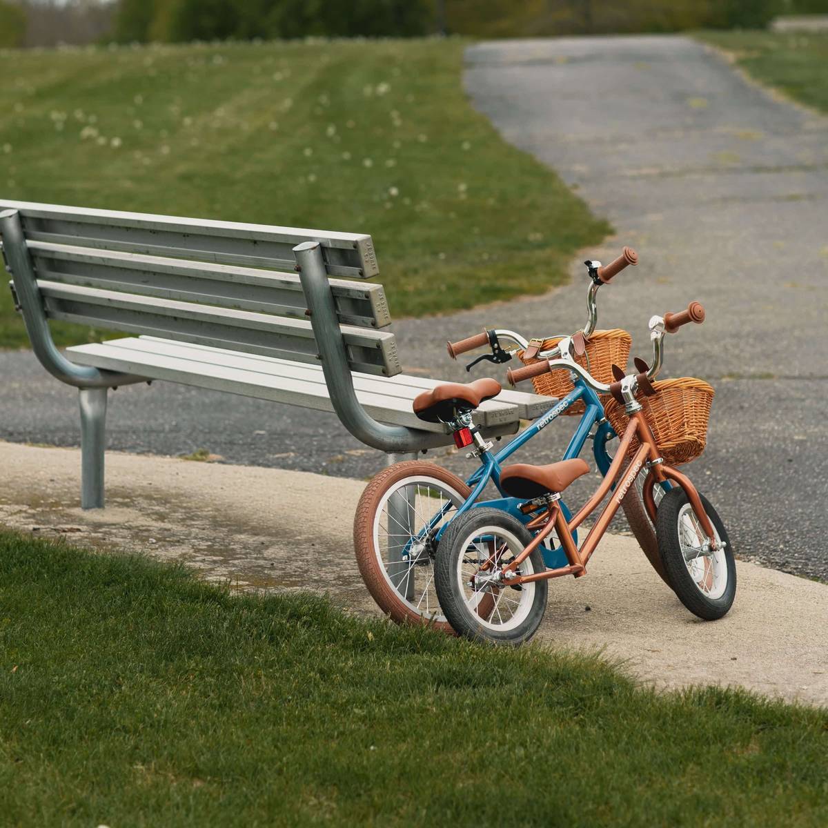 Two Retrospec kids bikes with wicker front baskets and brown leather-look grips parked side by side next to a park bench along a paved path.