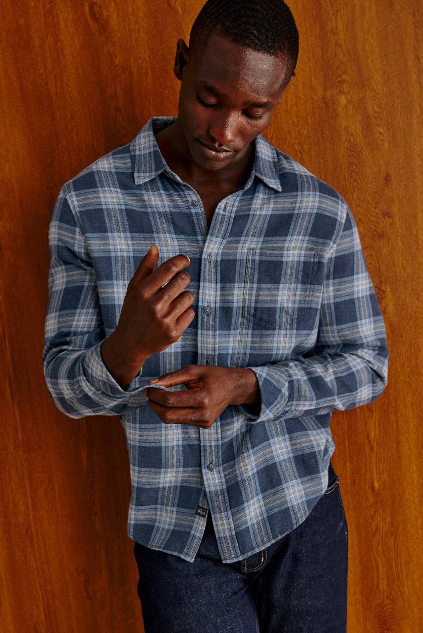 Man leaning against a warm wood-paneled wall wearing a long-sleeve blue plaid flannel shirt in navy, slate blue, and cream, styled with dark indigo jeans.