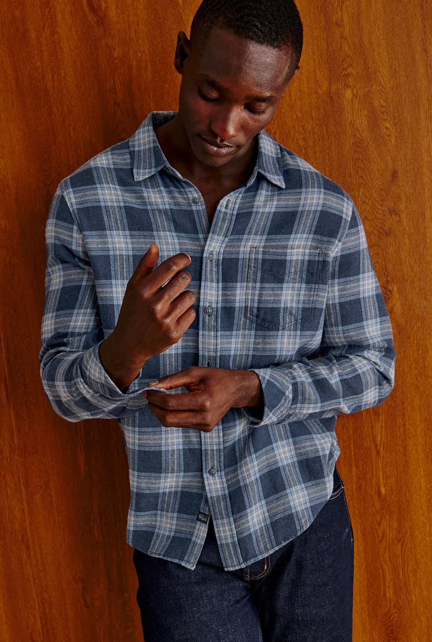 Man leaning against a warm wood-paneled wall wearing a long-sleeve blue plaid flannel shirt in navy, slate blue, and cream, styled with dark indigo jeans.