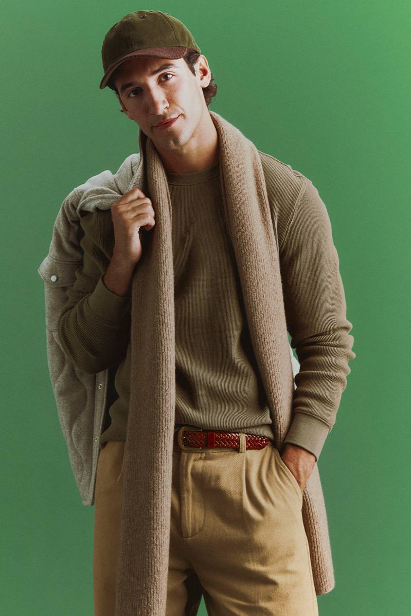 A man in an olive cap and olive waffle-knit top wears a tan knit scarf, khaki pleated pants, and a braided leather belt, with a quilted jacket slung over his shoulder against a green backdrop.