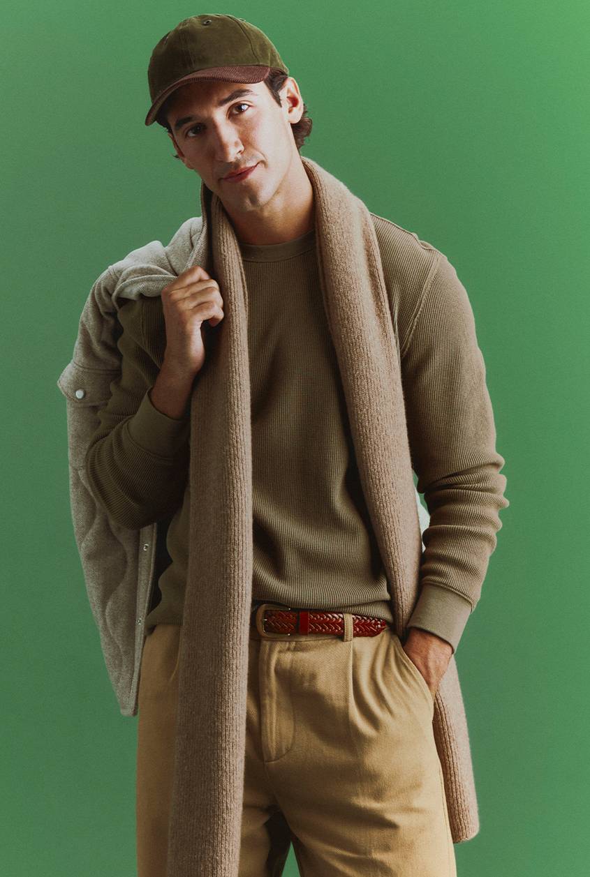 A man in an olive cap and olive waffle-knit top wears a tan knit scarf, khaki pleated pants, and a braided leather belt, with a quilted jacket slung over his shoulder against a green backdrop.