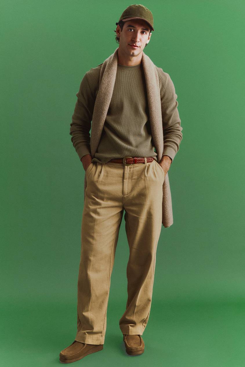 Man in an olive cap, olive waffle-knit top, khaki pleated trousers, and suede shoes, with a beige knit scarf draped over his shoulders, standing against a green backdrop.