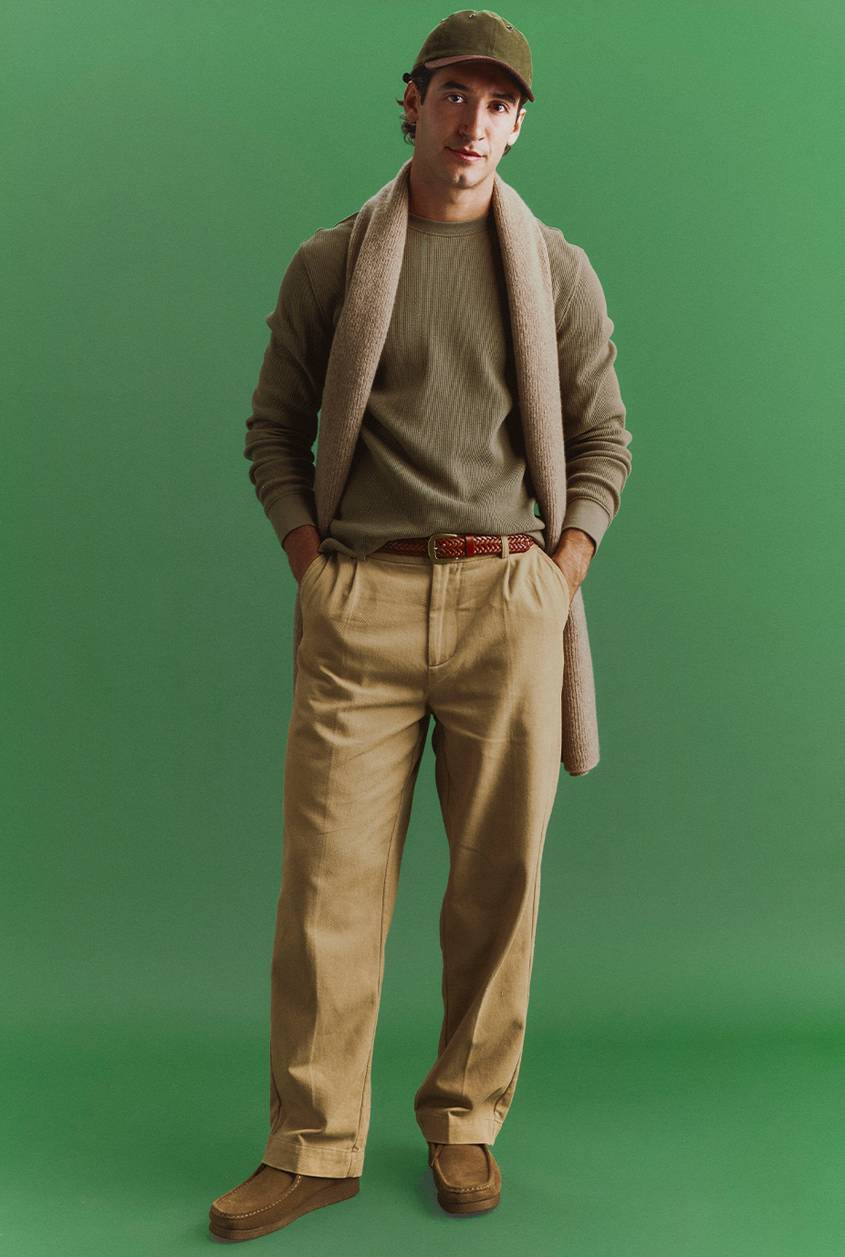 Man in an olive cap, olive waffle-knit top, khaki pleated trousers, and suede shoes, with a beige knit scarf draped over his shoulders, standing against a green backdrop.