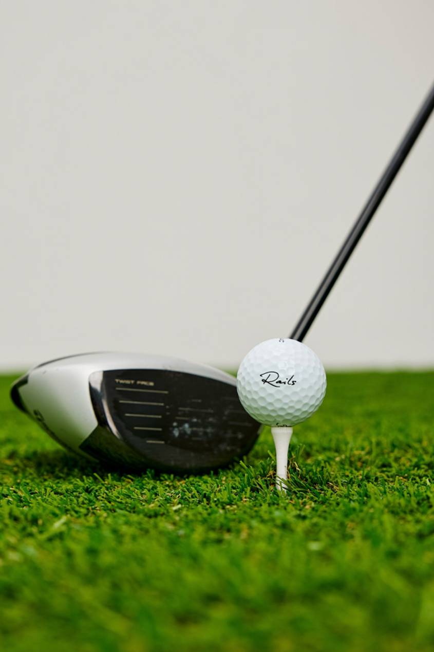 A close up shot of a golf club and a Rails exclusive golf ball displayed on a golf tee and green grass.