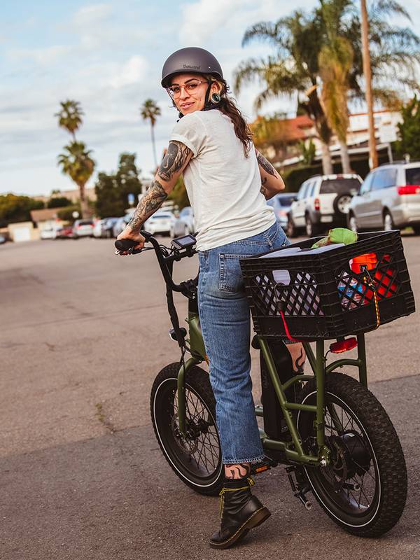 Image of a woman on the RadRunner 2 in Forest Green with a large basket on the back. She is wearing a helmet & has tattoos on her arm. 
