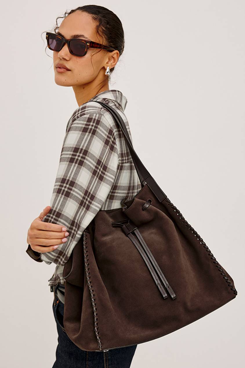 Woman wearing a gray plaid shirt and carrying a large brown suede bucket bag.