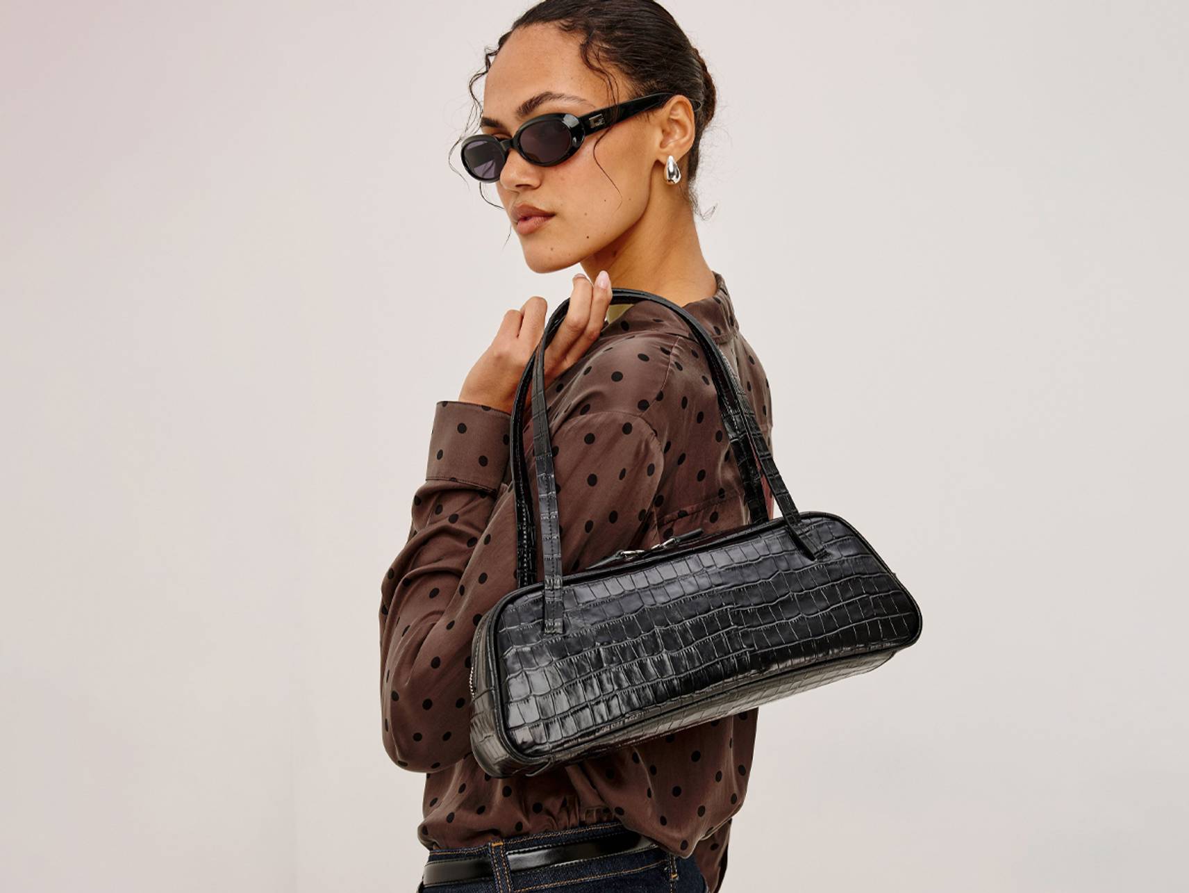 Woman wearing sunglasses and a brown polka-dot blouse, carrying a black croc-effect shoulder bag.