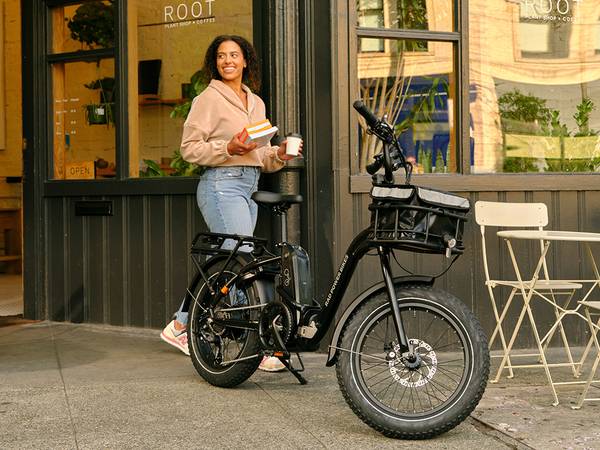 Woman walking out of store with a cup of coffee and books, walking towards her step-thru RadExpand folding electric bike