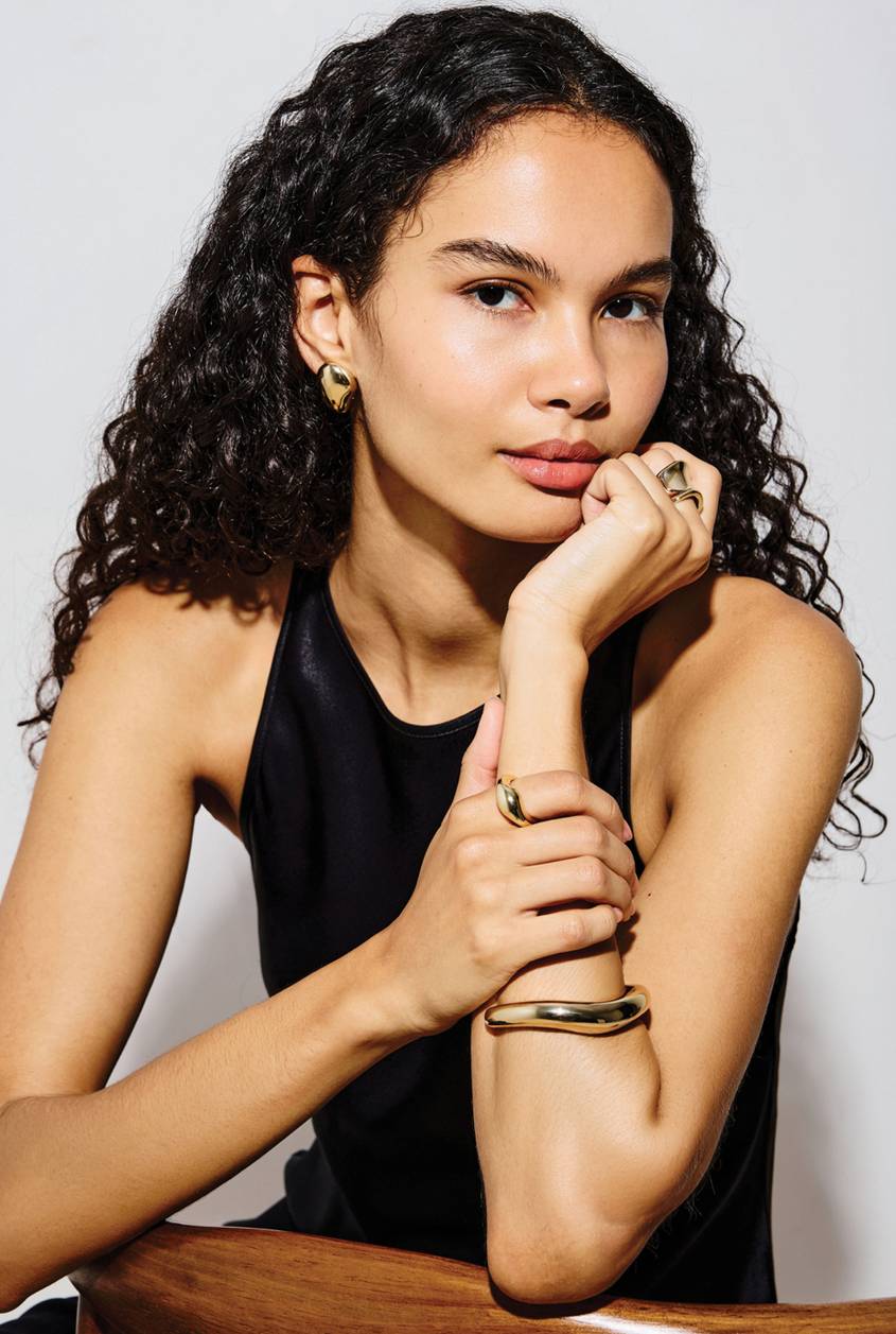 Model wearing a black sleeveless top and gold jewelry, including statement earrings, rings, and a cuff bracelet.