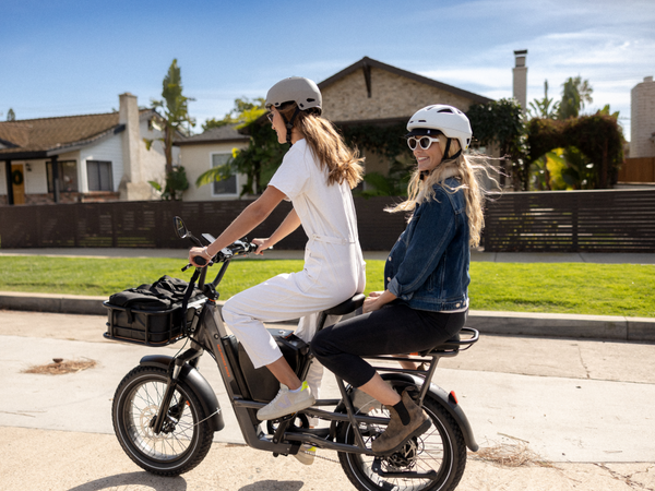 Two women riding a RadRunner 3 Plus electric utility bike, with the passenger seated on a passenger package seat.