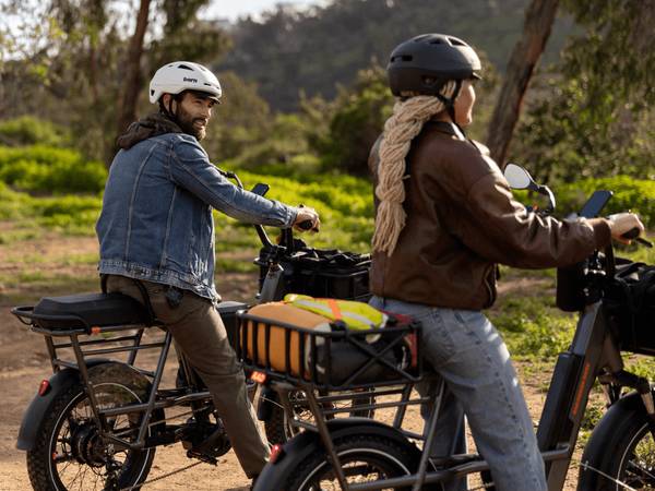 Two people each riding a RadRunner 3 Plus with helmets on. One person is a man with a jean jacket, the other is a woman with a long braid & leather jacket.