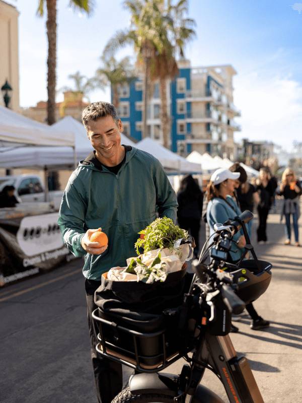Image of a man at a farmers market adding an orange to his front-mounted basket bag. He's smiling & wearing a green sweatshirt.  