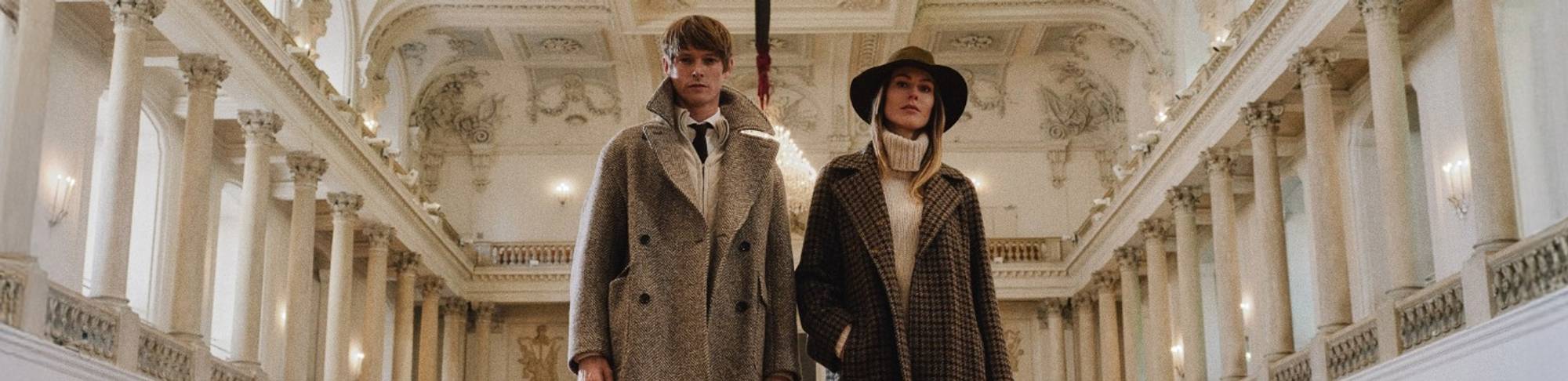 Man and woman standing side by side in an ornate hall with high decorated ceilings, both wearing long patterned coats; the woman accessorised with a wide-brim hat and turtleneck sweater