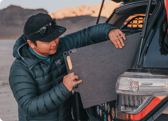 A person in a puffy jacket attaches a gray panel to the back of a vehicle in a desert setting.