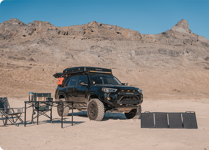 A black SUV parked on a sandy area with mountains in the background, accompanied by camping chairs and solar panels.