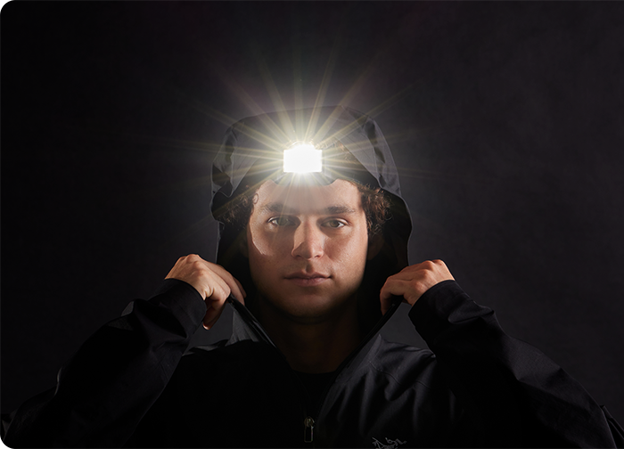 A person wearing a black jacket with a headlamp, illuminated against a dark background.