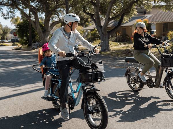 An image of a family riding their Rad Power Bikes.   On the left, a man is riding the RadWagon 5 Plus in Metallic blue with a child on the back. On the right, a woman is riding a RadRunner 3 Plus. Everyone is wearing a helmet. 