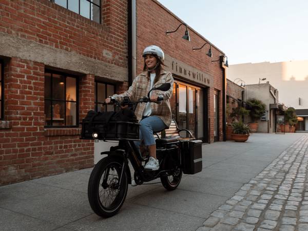 An image of a woman riding a RadWagon 5 on a city sidewalk. The bike is outfitted with a large front-mounted basket & rear-mounted panniers. She is wearing a helmet. 