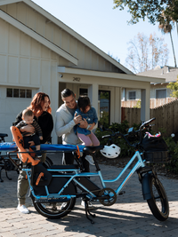 An image of a family in the the driveway of their house with the RadWagon 5 Plus in Metallic blue. The ebike is kitted with a Thule Yepp Child seat, caboose, large front-mounted basket & large basket liner. 