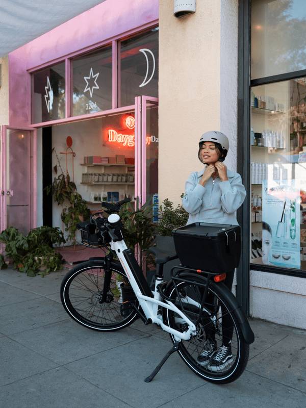 Person putting on their helmet standing next to a Radster Road electric commuter bike outfitted with accessories, including a hardshell locking box, front basket, mirror and phone mount.