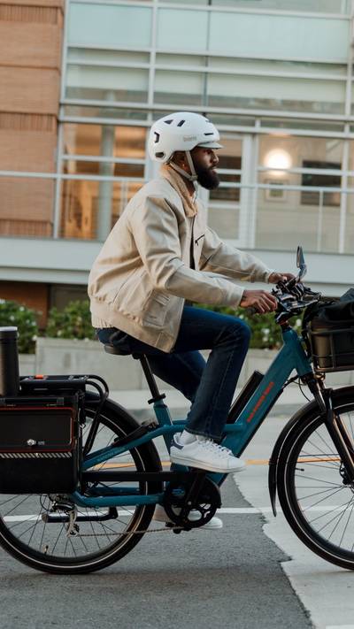 Man riding on the street on a blue Radster Road electric commuter bike with a front basket and hardshell pannier. 