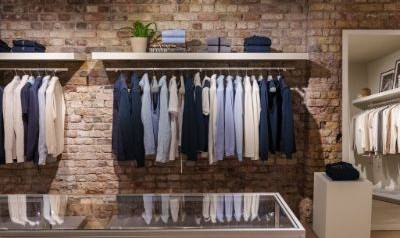 Clothing display against an exposed brick wall with neatly hung shirts and knitwear in neutral and blue tones, a glass-topped display table in the foreground, and folded garments on side shelves, inside the Sloane Street store