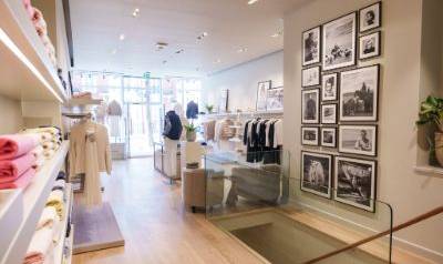 N.Peal Sloane Street store interior with folded knitwear on side shelves, clothing racks in the centre, a wall display of framed black-and-white photographs, and large windows letting in natural light at the entrance