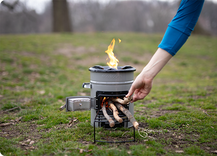 A person adds sticks to a portable stove with flames, set on grass in a park.
