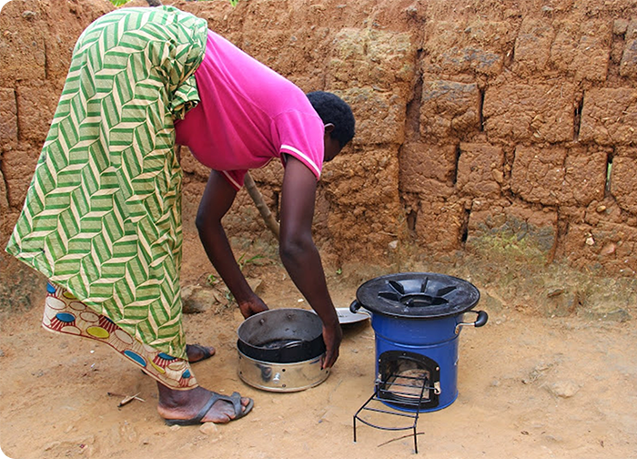 A person in a colorful skirt is preparing to cook with a blue stove outdoors, next to a mud wall.