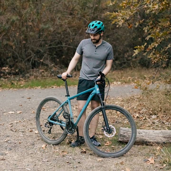 A person stands beside a teal mountain bike on a gravel path surrounded by autumn foliage, wearing a Rowan Mountain Bike Helmet and casual cycling attire.