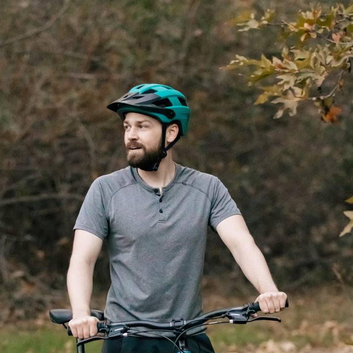 A person in a gray shirt and teal Rowan Mountain Bike Helmet stands beside a bicycle, surrounded by autumn foliage in a wooded area.