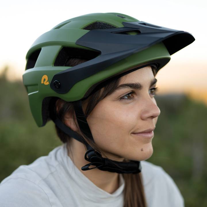A person wears a green Rowan Mountain Bike Helmet with a black visor, set against a blurred natural background during sunset.