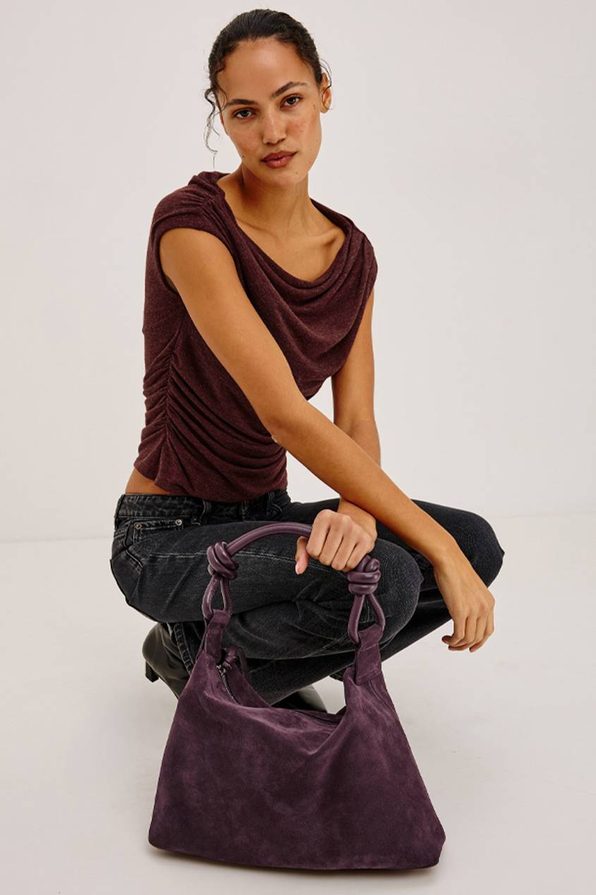 Woman wearing a burgundy draped top and dark jeans, holding a purple suede handbag.