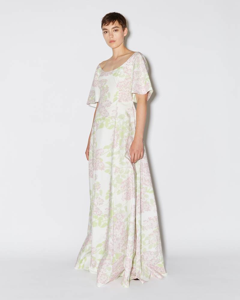 Bernadette Antwerp dress Maude is made from cotton, features short sleeves, an accentuated waistline, and a boat-neckline that flows into a grand floor-length gown. Evening wear dress adorned with Lilacs floral print in colors Pink and White. 
