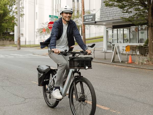 Man riding the RadKick 7-Speed in Arctic Blue down a city street with a front-mounted basket & rear-mounted cargo bags. He is wearing a helmet. 