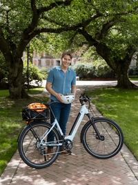 A male college student standing next to the RadKick 7 Speed in Arctic Blue. He is holding a helmet & his bike is equipped with a rear-mounted large basket. 