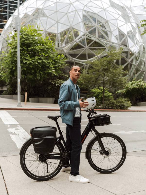 Man standing over the RadKick Belt Drive in Midnight Blue with rear-mounted pannier bags & front-mounted large basket. He is holding a helmet. 